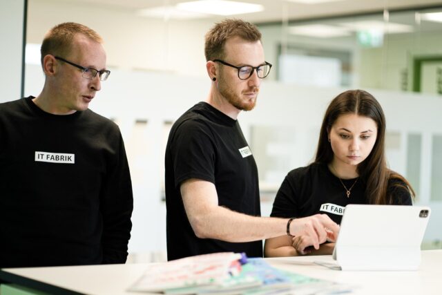 Drei Fachkräfte der IT FABRIK im Gespräch vor einem Tablet; sie tragen schwarze Shirts mit dem Firmenlogo und arbeiten gemeinsam an einer Lösung in einem modernen Büro.